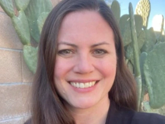 Kelley wears a black blazer and dark maroon blouse. She smiles in front of a brick wall and nopal cactus.