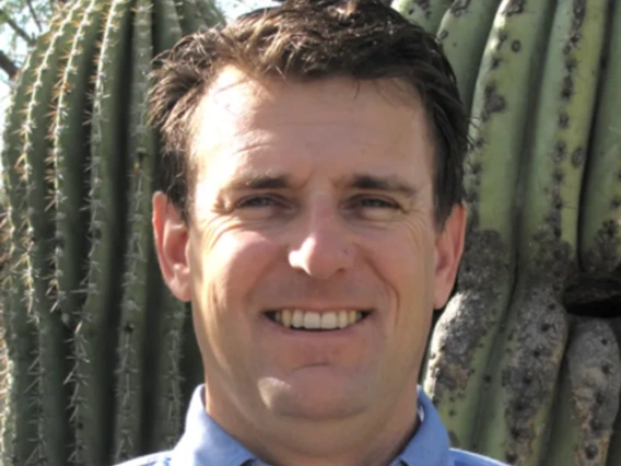 Kevin Bonine wears a blue button-up collared shirt and smiles at the camera. He stands in front of a Saguaro cactus.