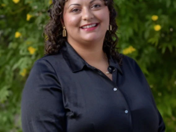 Tori Nardinelli wears a dark button-up shirt as she smiles in front of a green leafy backdrop with yellow flowers.