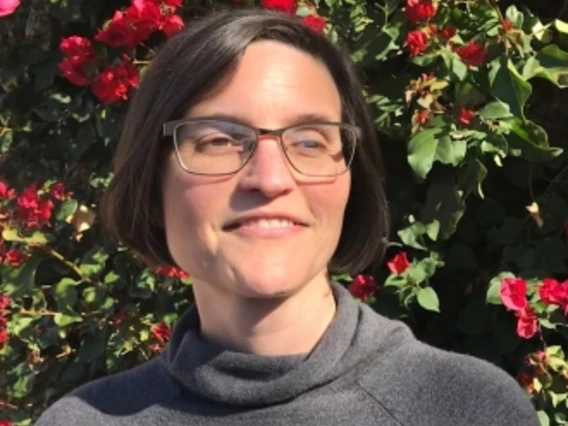 Kay wears glasses, a warm grey shirt, and short hair as she stands in front of a blooming bougainvillea bush on a sunny day.